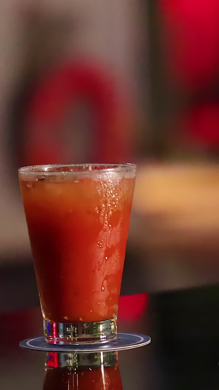 Una mano versa whisky in un bicchiere da cocktail alto e rosso su un bancone da bar lucido, illuminazione calda