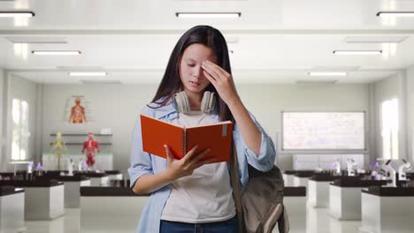 asian teen girl student with a backpack reading book and having a headache while standing in science laboratory