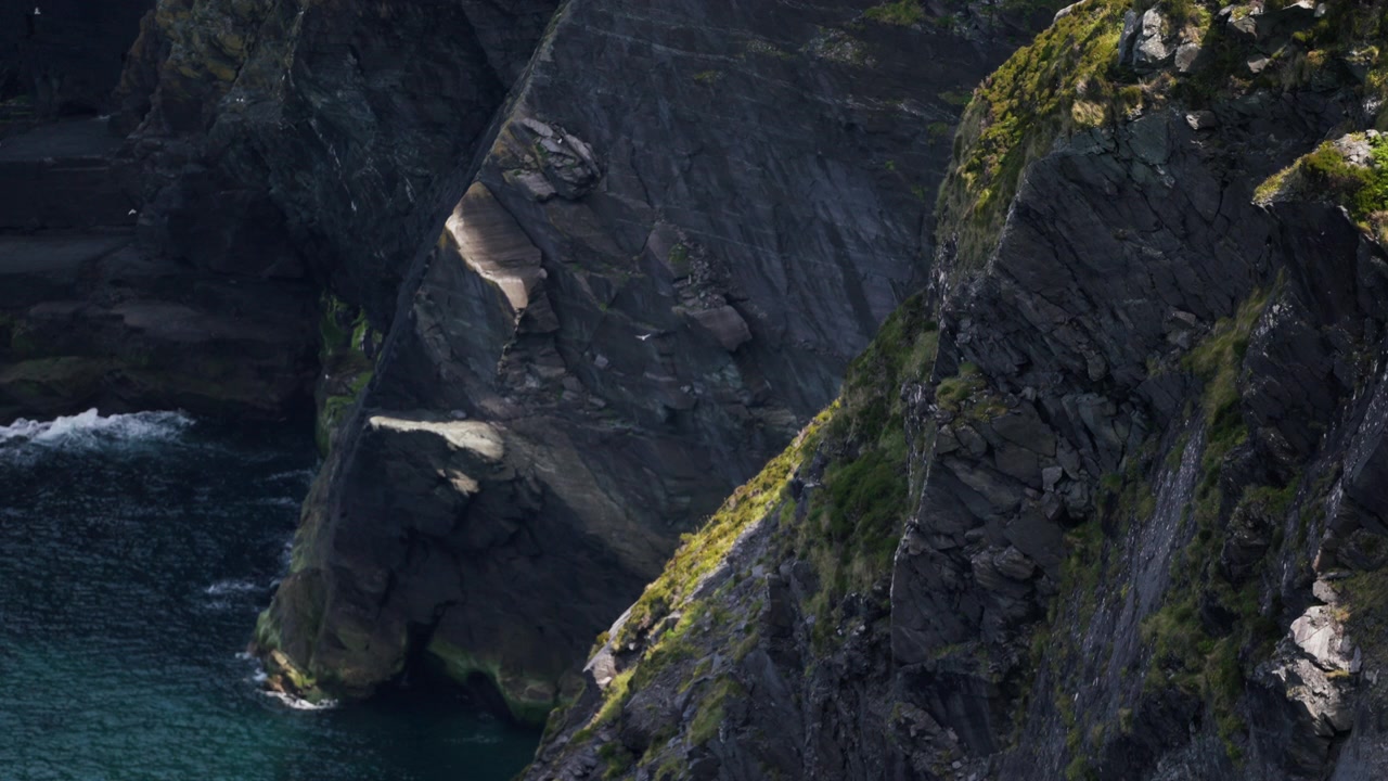 Rocky cliffs rise steeply from the ocean with waves crashing below and a seabird flying near the rugged coastline.