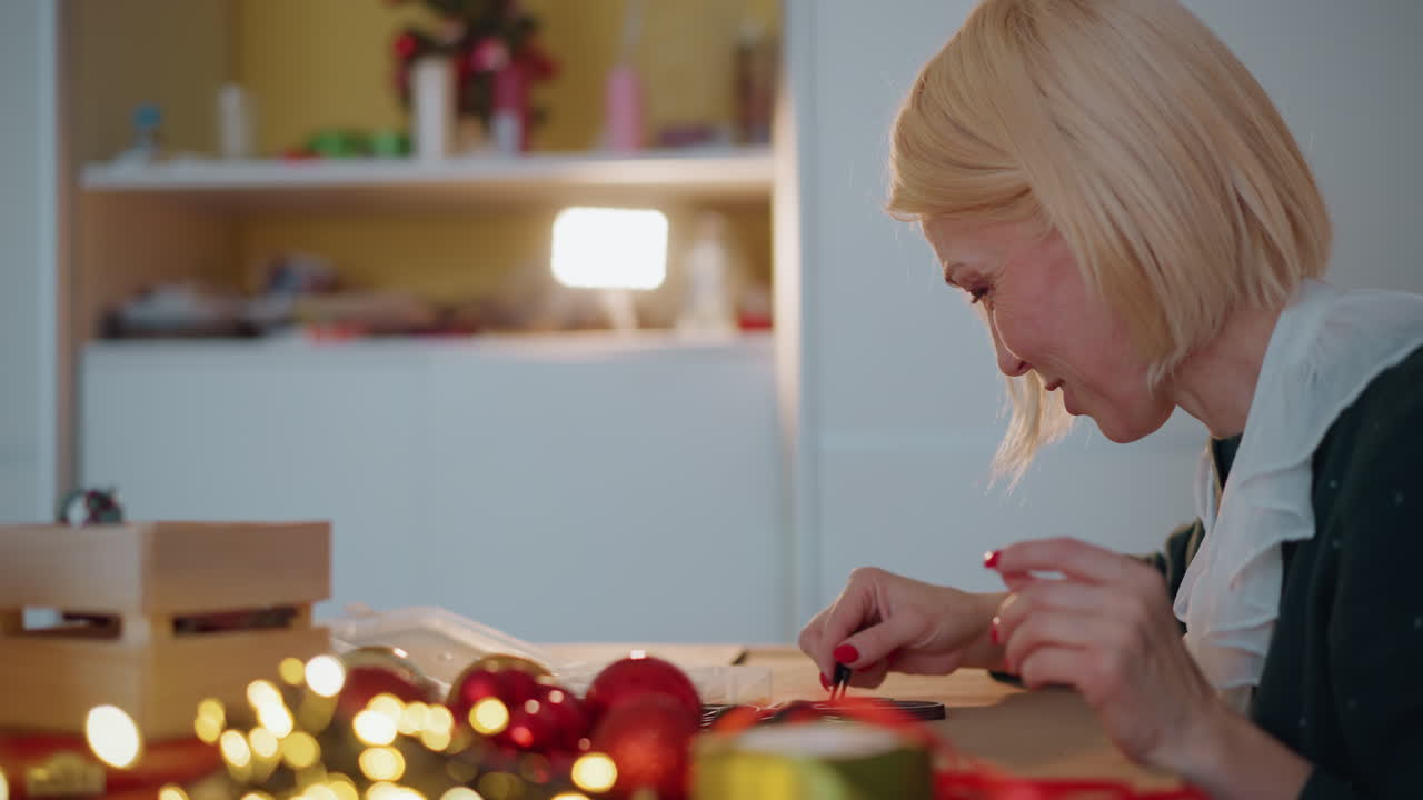 Blonde woman wrapping christmas gifts at table, cutting ribbon, arranging baubles and boxes, candle glow and fairy lights foreground, focused concentration, craft tools and festive decor, holiday