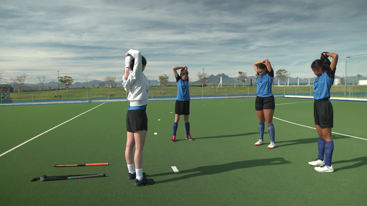 Female hockey players stretching on field, preparing for practice under clear sky
