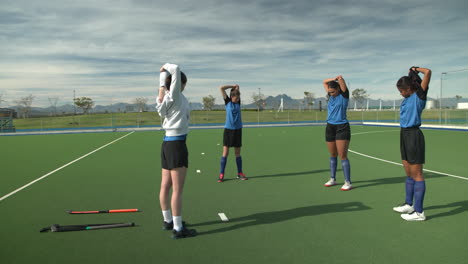 female hockey players stretching on field, preparing for practice under clear sky