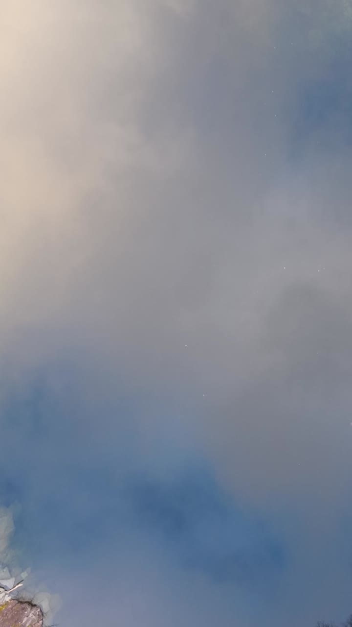 Vertical drone top down ascent above a quarry lake that mirrors blue sky and drifting clouds, gradually revealing the rocky edges and towering slate walls in a hypnotic view