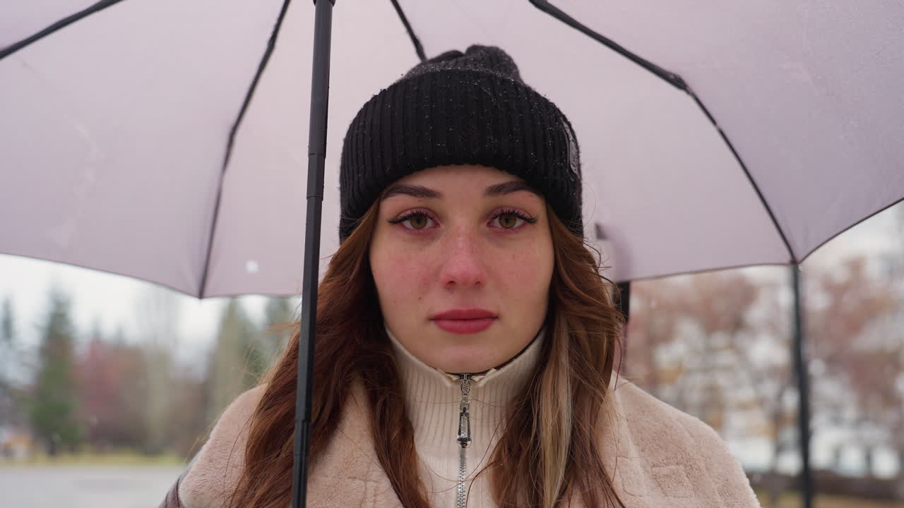 Lady with serious expression standing still, holding umbrella, wearing black knit cap and brown shearling jacket, under cold overcast sky with light snowfall
