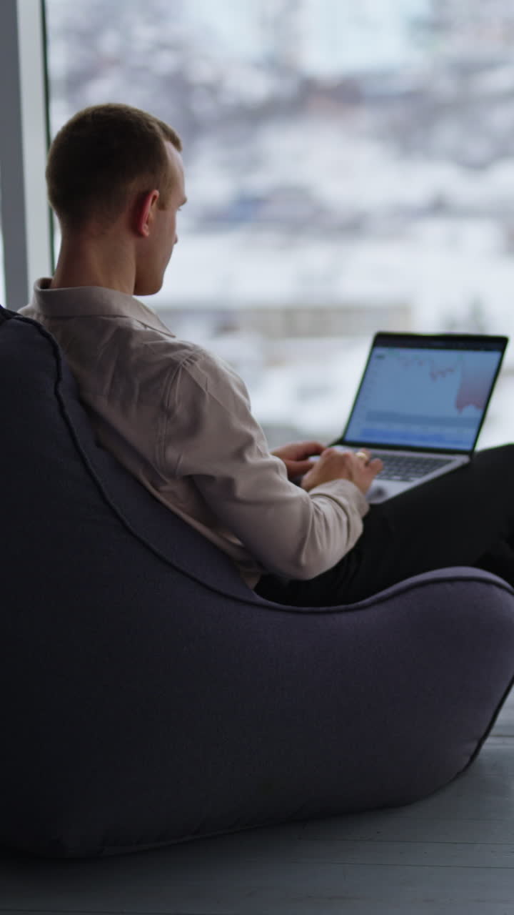 Working man sits with his back to the camera typing on his laptop. Male employee building charts on computer. City panorama in blur at backdrop. Vertical video