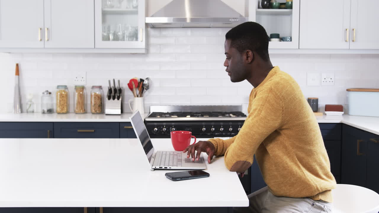 Man working on laptop in kitchen with red mug and smartphone nearby