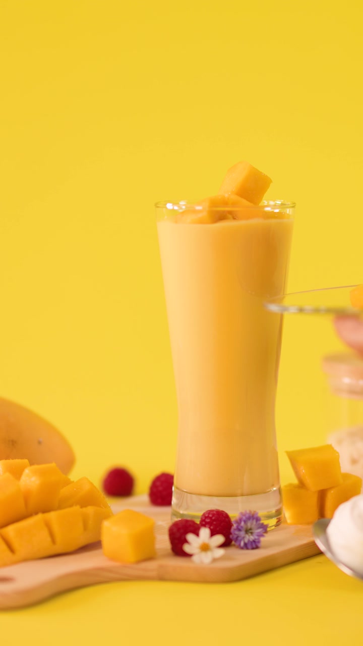 Una mano aggiunge cubetti di mango a un alto bicchiere da frullato su uno sfondo da studio giallo brillante, telecamera fissa.