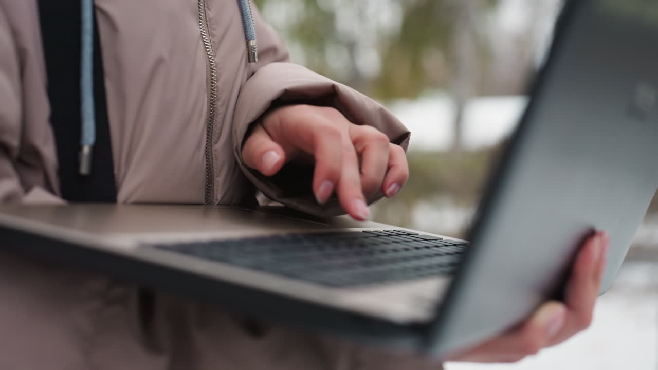 Close-up of light-skinned female hand typing on black laptop while holding it with other hand, wearing winter jacket outdoors, with softly blurred light and natural background creating calm atmosphere