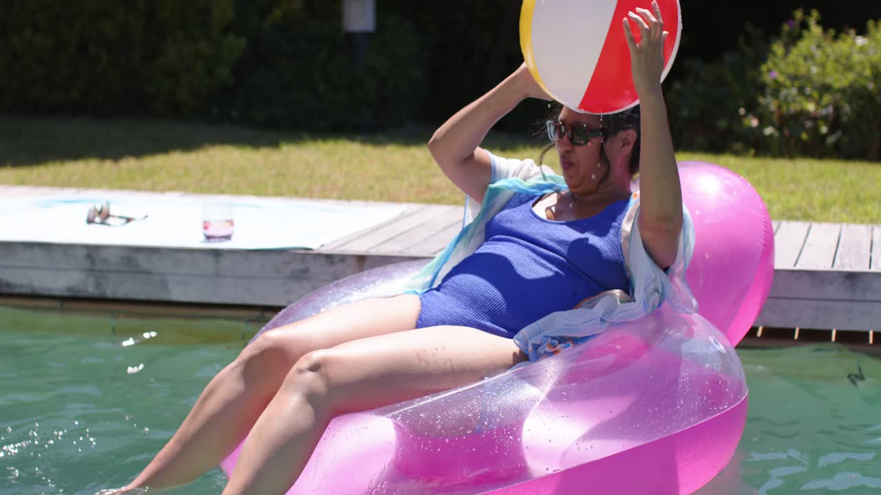 Tossing woman on pink ring hurling beach ball over home pool, friends in swimwear catching, playing