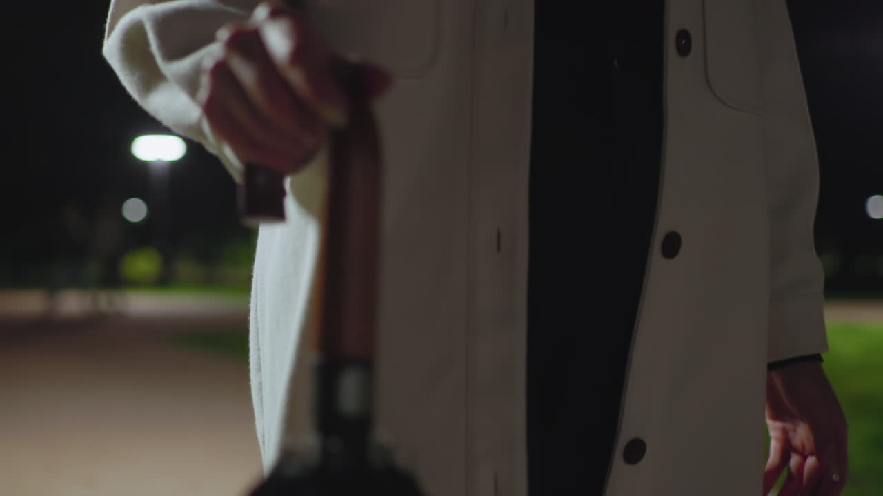 Close up of person wearing white coat walking at night holding umbrella handle, background featuring blurry green grass, glowing streetlights, and moving figure with soft bokeh lights