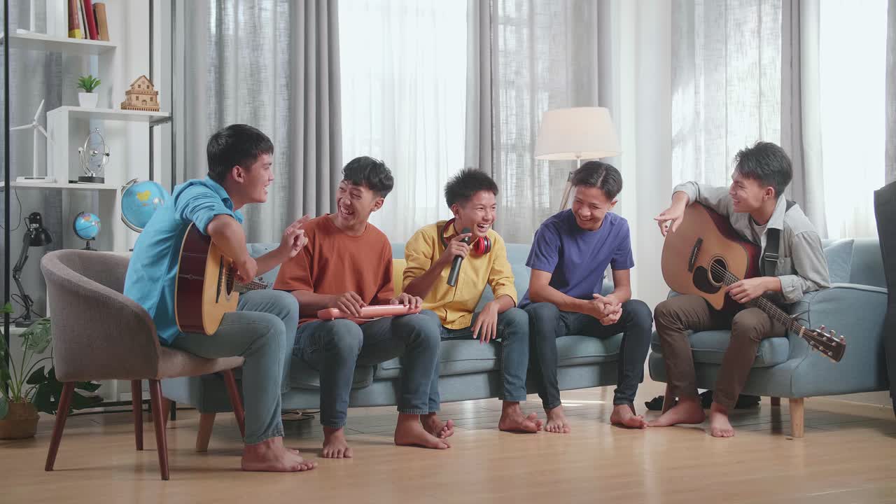 마이크와 악기를 가진 젊은 아시아 친구 그룹이 집에서 소파에 앉아서 말하고 웃고 있습니다.
