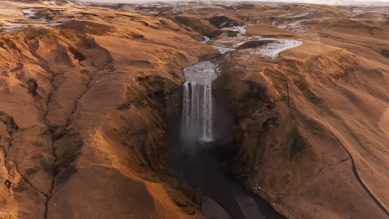 Drone flies slowly toward majestic Skógafoss waterfall in Iceland’s winter, cinematic aerial view of nature, river and snowy terrain.