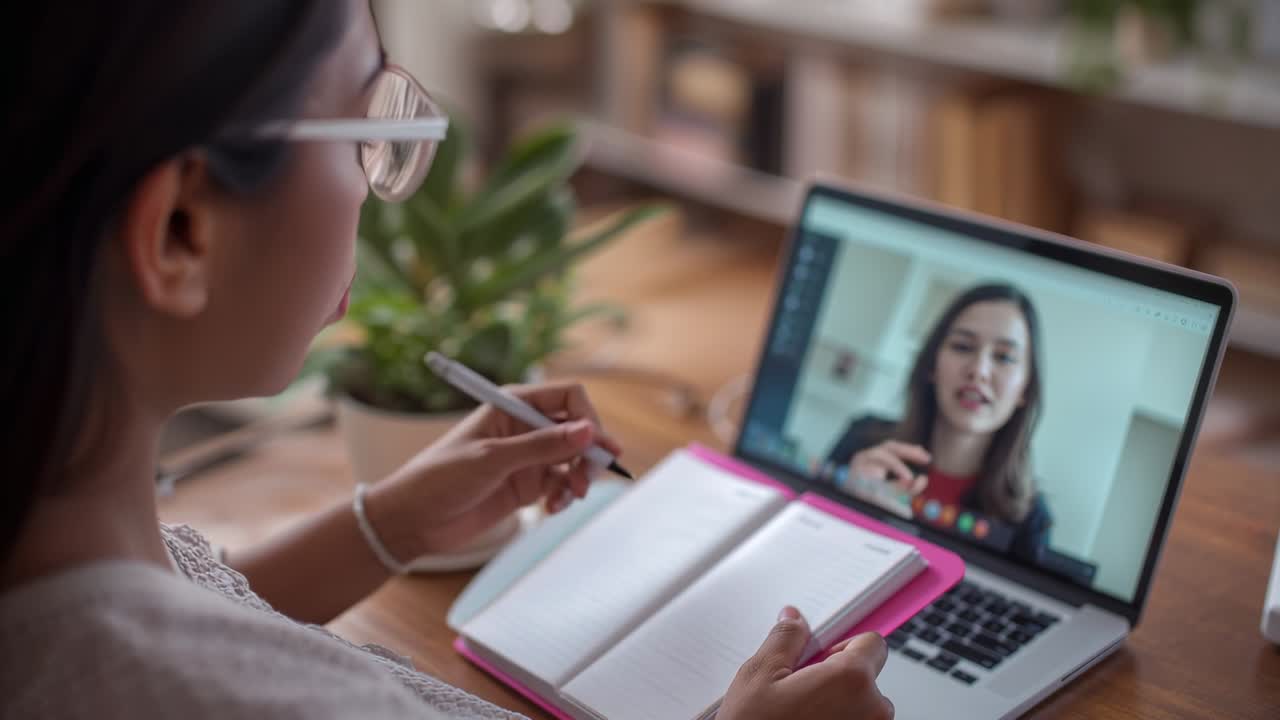 Woman jotting with pen as colleague speaking on laptop in home office, pink notebook, copy space