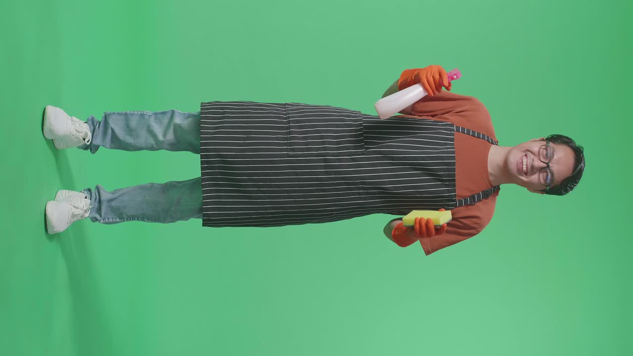 녹색 스크린 스튜디오에 서 있는 동안 스프레이와 부 스폰지를 바라보는 아프론을 입은 미소 짓는 아시아 남성 집주인