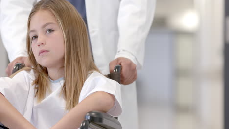 diverse male doctor pushing girl patient in wheelchair in hospital, copy space, slow motion