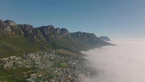 Amazing-panoramic-aerial-descending-footage-of-rocky-mountain-ridge-rising-over-landscape-flooded-by-coastal-fog.-Cape-Town,-South-Africa