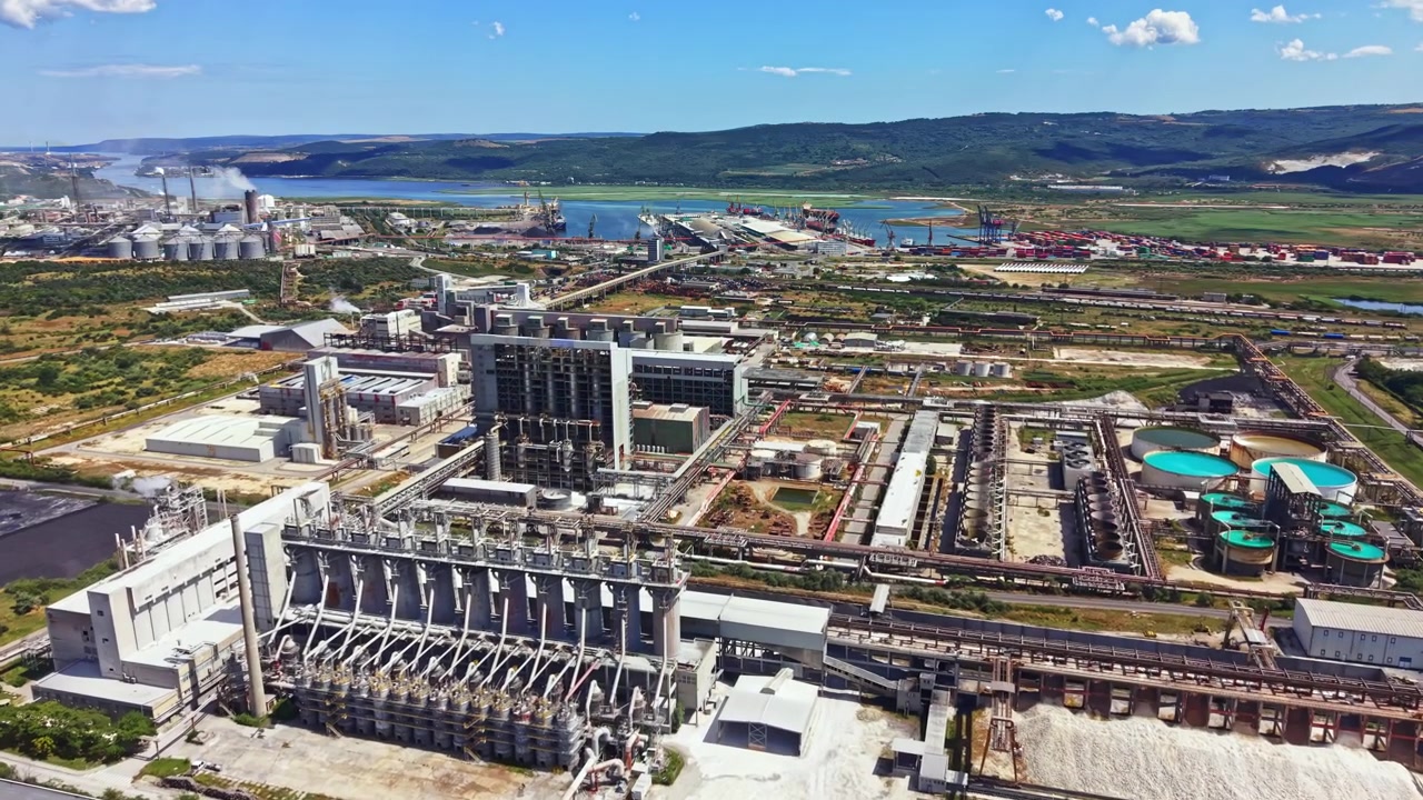 Sito industriale con edifici e strutture multiple situato vicino a un fiume, con un cielo sereno e colline distanti sullo sfondo