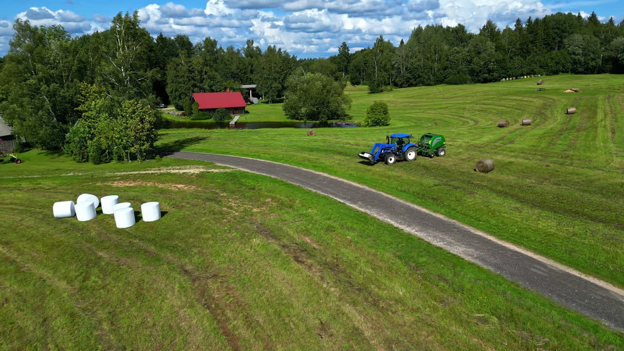 A tractor drives through a lush green countryside with bales scattered across open fields