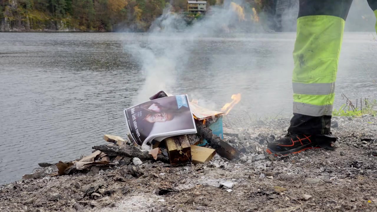 Bonfire burning advertising leaflets by the fjord, with a person adding more unwanted flyers