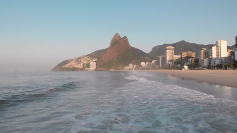 Low-aerial-view-above-waves-coming-in-on-coastal-city-beach-of-Rio-de-Janeiro-during-early-morning-golden-hour