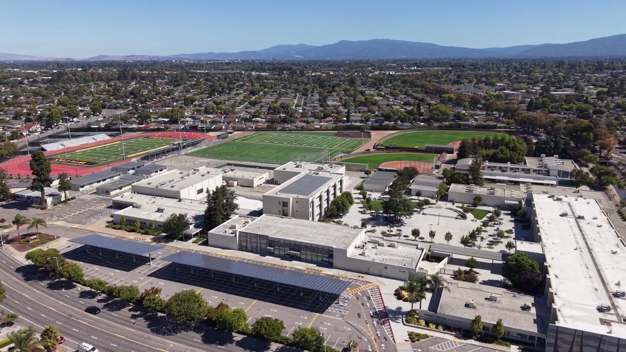Orbiting aerial perspective of a modern high school campus in Santa Clara, California, featuring sports fields, solar panels on carports, and surrounding suburban neighborhoods on a clear day