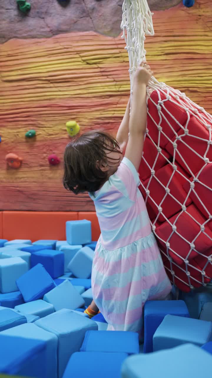 Happy young girl in a blue foam block pit reaching up to hold a large red sack of suspended foam blocks at an indoor playground