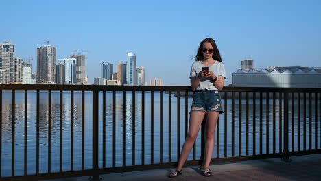 beautiful girl with long hair in sunglasses using smartphone app at sunset river quay near.