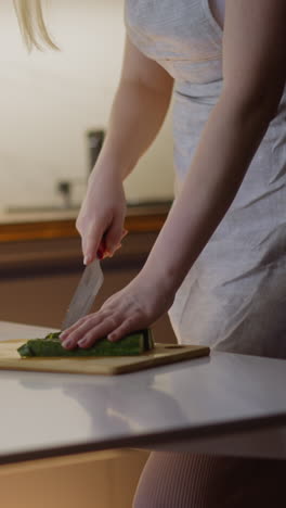 housewife in grey apron cuts juicy cucumber on wooden board to make healthy salad for supper on table in light kitchen close view slow motion