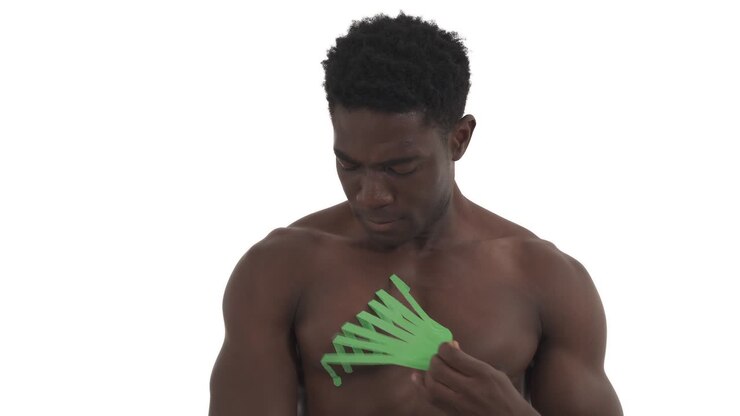 Primo piano di un atleta maschio che rimuove il taping kinesiologico verde dalla spalla e compie movimenti circolari con la spalla. Trattamento della spalla infortunata tramite taping kinesiologico. Isolato su sfondo bianco.