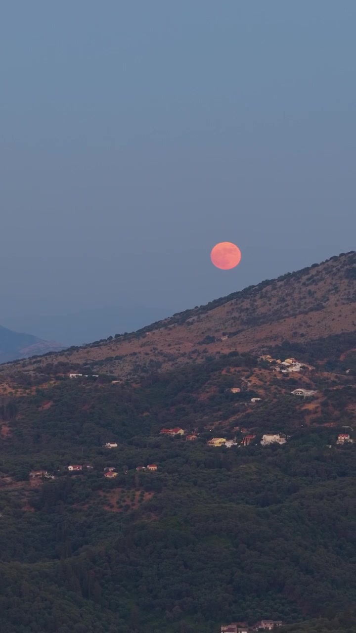 A stunning time-lapse capturing a large, glowing orange full moon rising majestically from behind the mountainous ridges of Corfu Island, Greece. The early evening Mediterranean landscape showcases ru