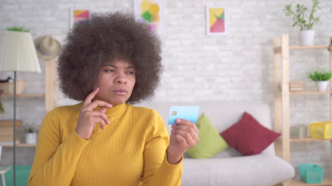 Beautiful african american woman with an afro hairstyle thoughtful and serious looking at a Bank card in his hands in a modern apartment