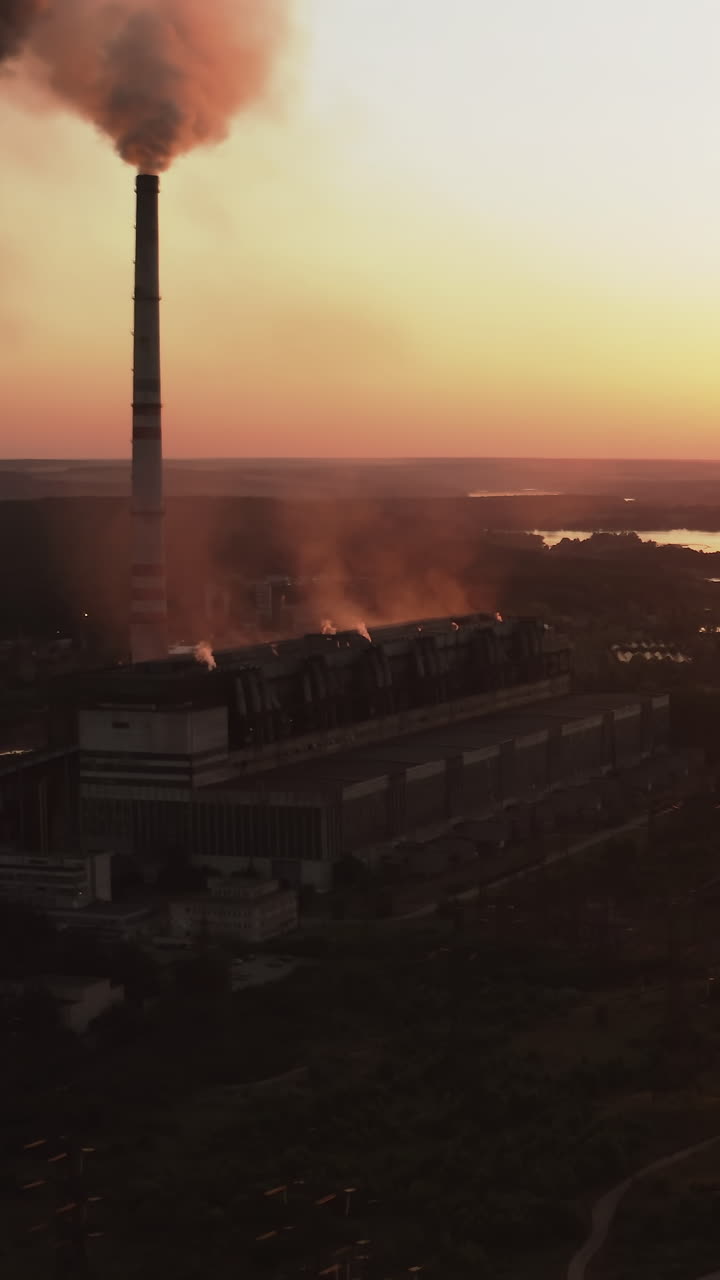 Silhouette of a factory near the river at sunset. Dark emissions from chimneys exhaust into the atmosphere against the orange setting sun. Ecology in danger. Vertical video
