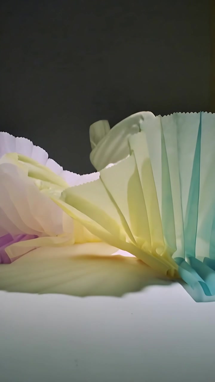 Video verticale: Carta plissettata che ondeggia per una brezza fuori campo su un tavolo, rivelando tonalità pastello.