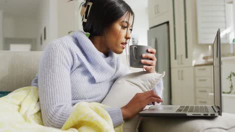 video of happy biracial woman in hijab sitting on sofa with coffee and using laptop