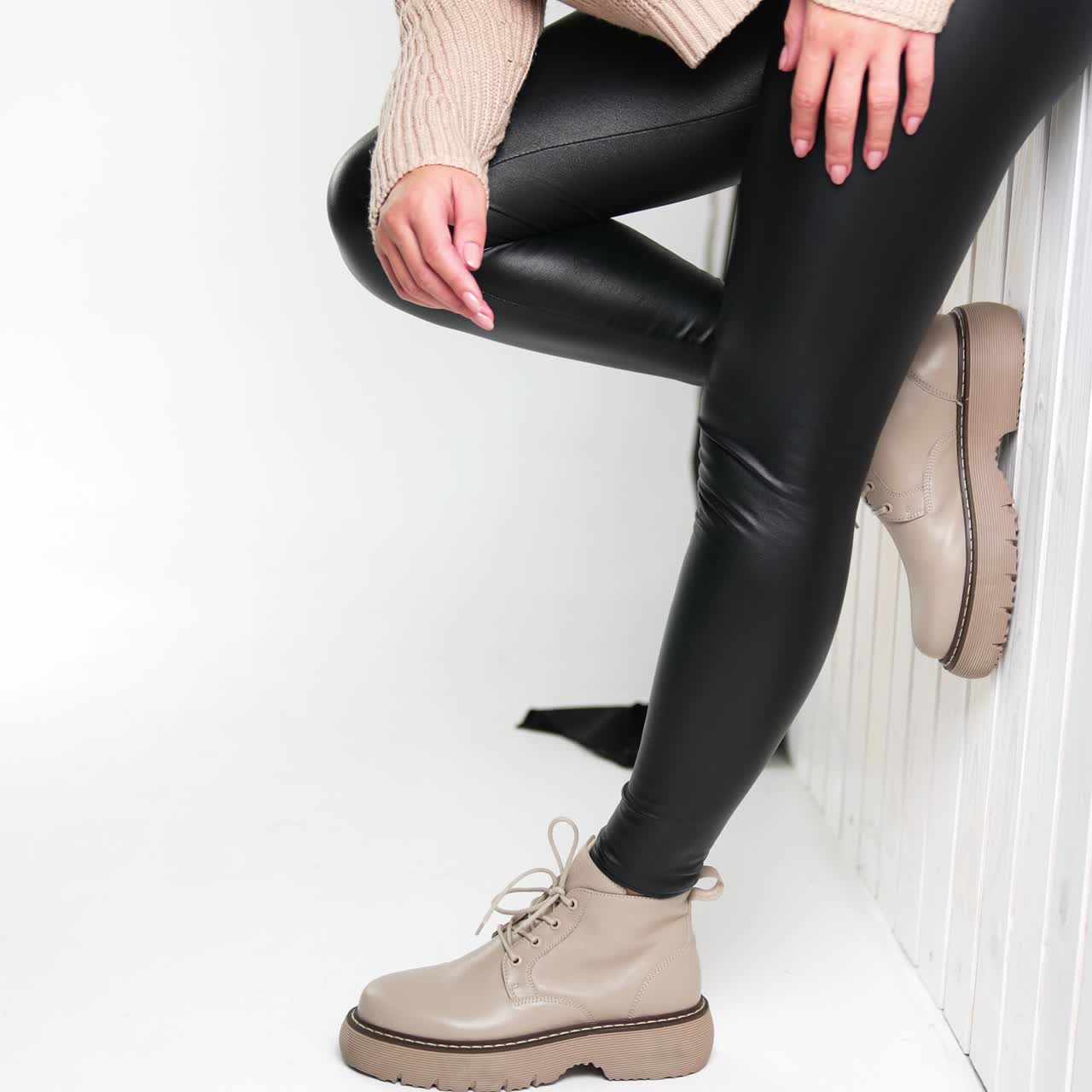 Model wearing black leather leggings beige sweater and boots stands leaned to the wall. Female demonstrating modern fashionable look