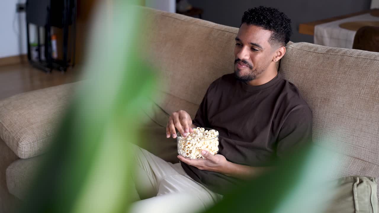 hombre étnico feliz comiendo palomitas de maíz y viendo televisión en el sofá