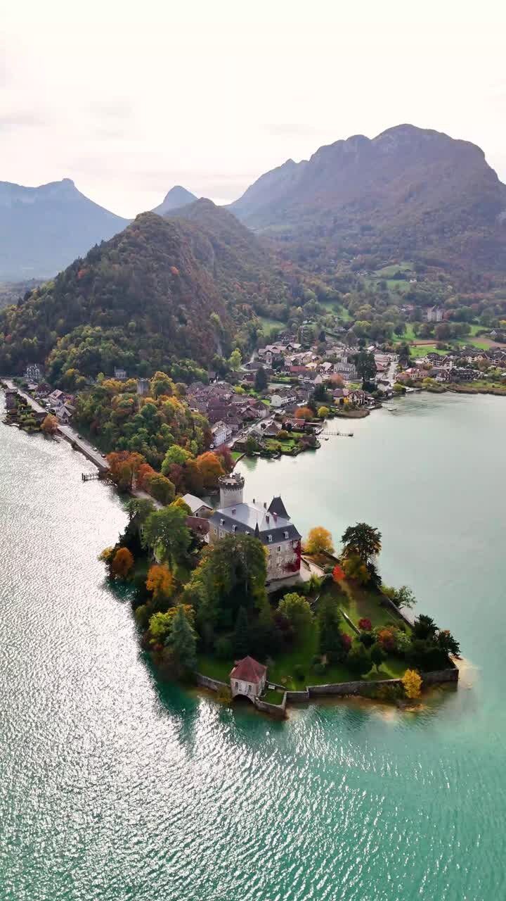 Il castello di Duingt con la sua torre, i giardini e il muro sul lago, poi il villaggio di Duingt e le montagne prealpine circostanti in Alta Savoia, Auvergne-Rhône-Alpes, Francia orientale, ripresa panoramica verticale con drone