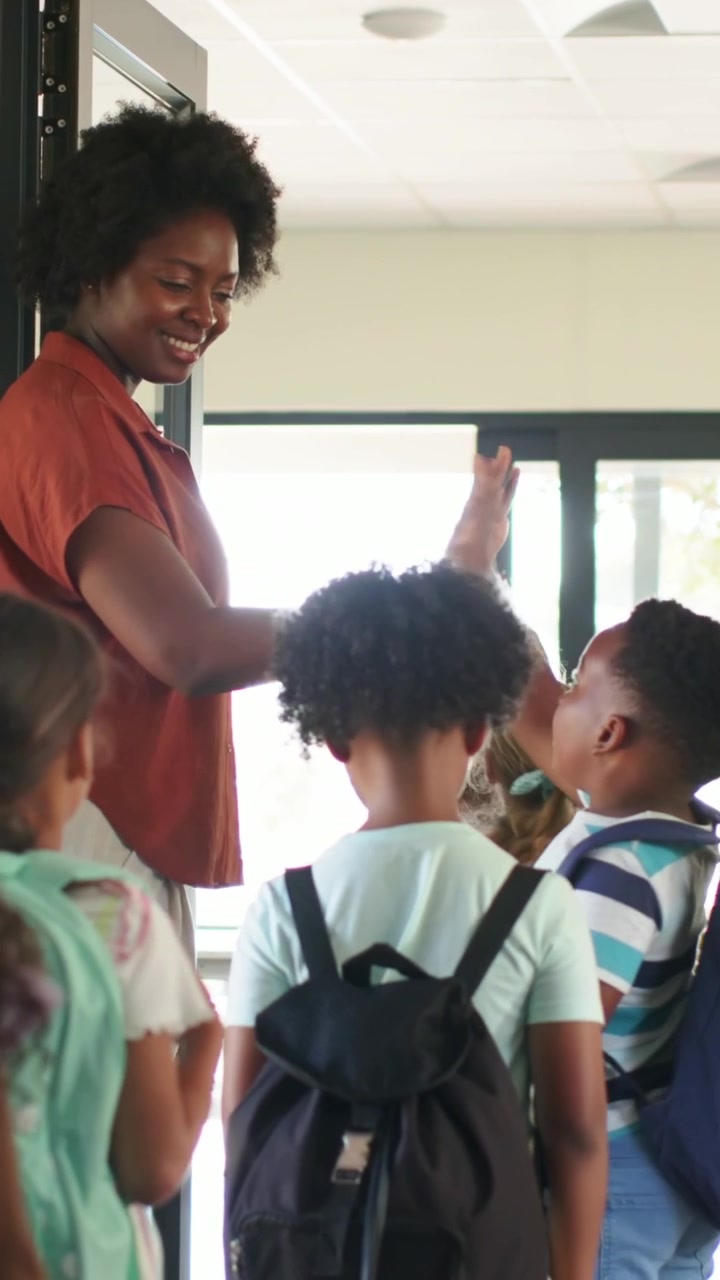 Video verticale: Insegnante che accoglie gli studenti in arrivo con il cinque, guidando gli zaini nella sala della scuola.