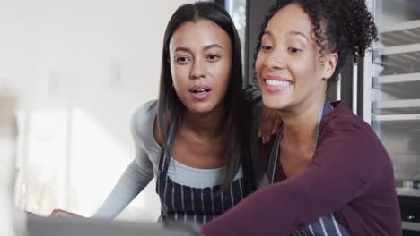 Happy-biracial-lesbian-couple-in-aprons-preparing-food-and-using-tablet-in-kitchen,-in-slow-motion