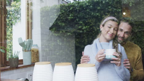 Happy-diverse-couple-embracing-and-looking-through-window-at-home,-in-slow-motion,-copy-space