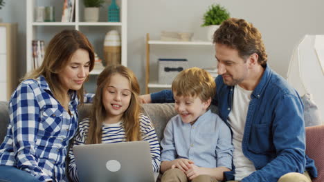 Padres-Jóvenes-Alegres-Con-Una-Hija-Pequeña-Y-Un-Hijo-Sentados-En-El-Sofá-De-La-Sala-De-Estar-Y-Hablando-Mientras-Tienen-Videochat-En-La-Computadora-Portátil