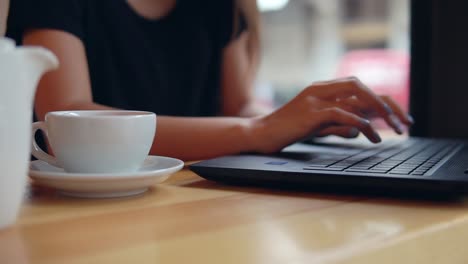 Close-Up-view-of-woman's-hands-typing-on-the-laptop's-keyboard-in-the-coffee-shot-during-a-coffee-break.-White-cup-with-saucer