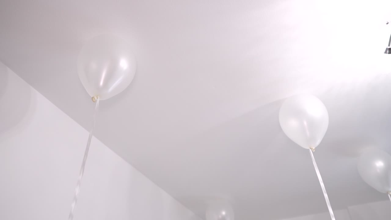 Balloons hang from the ceiling in a room decorated for a celebration. The setting features white colors and a soft light, creating a simple festive atmosphere