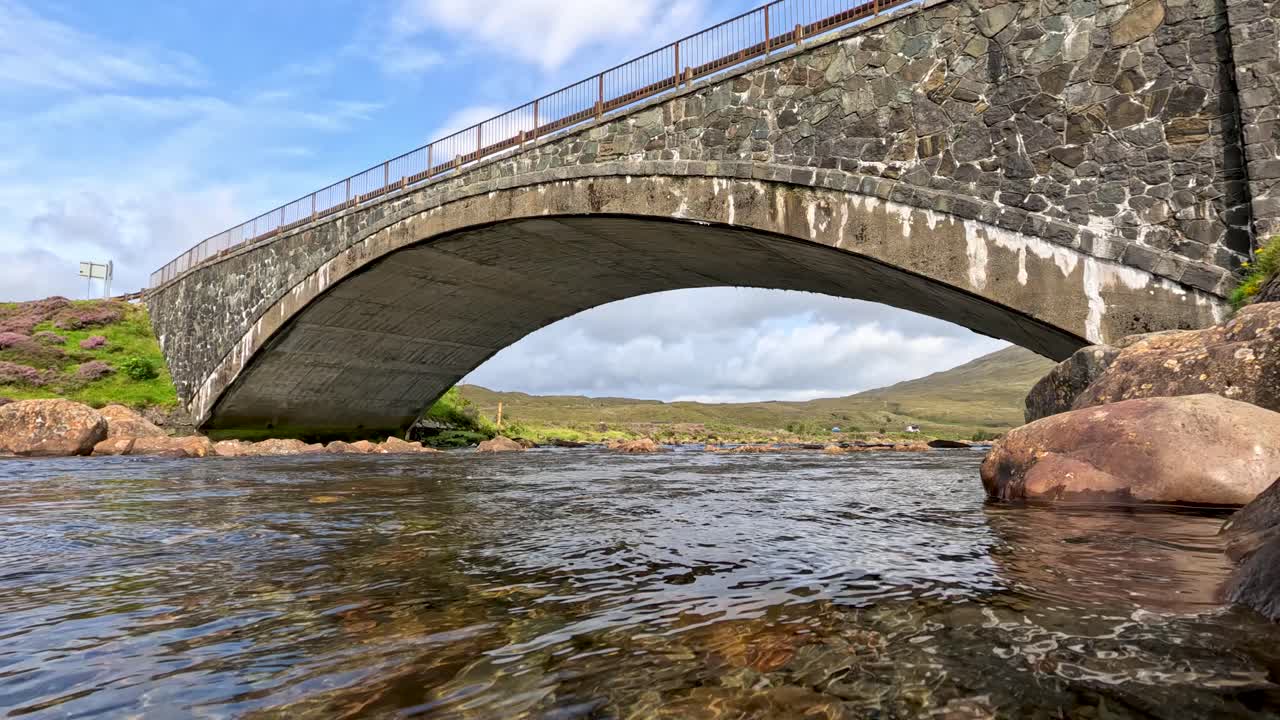 Low-angle view of a stone arch bridge crossing a clear stream, with gentle camera movement and natural daylight highlighting the rugged Highland landscape