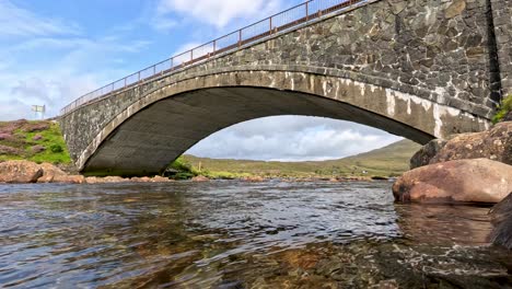 low-angle view of a stone arch bridge crossing a clear stream, with gentle camera movement and natural daylight highlighting the rugged highland landscape