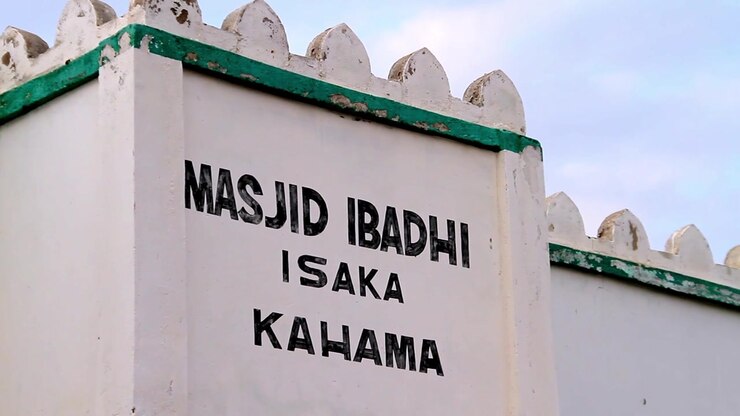 Üzerine siyah renkte "Masjid Ibadhi Isaka Kahama" yazısı yazılmış, dekoratif mazgallı üst kenarlara sahip beyaz bir duvarın yakın çekim görünümü.