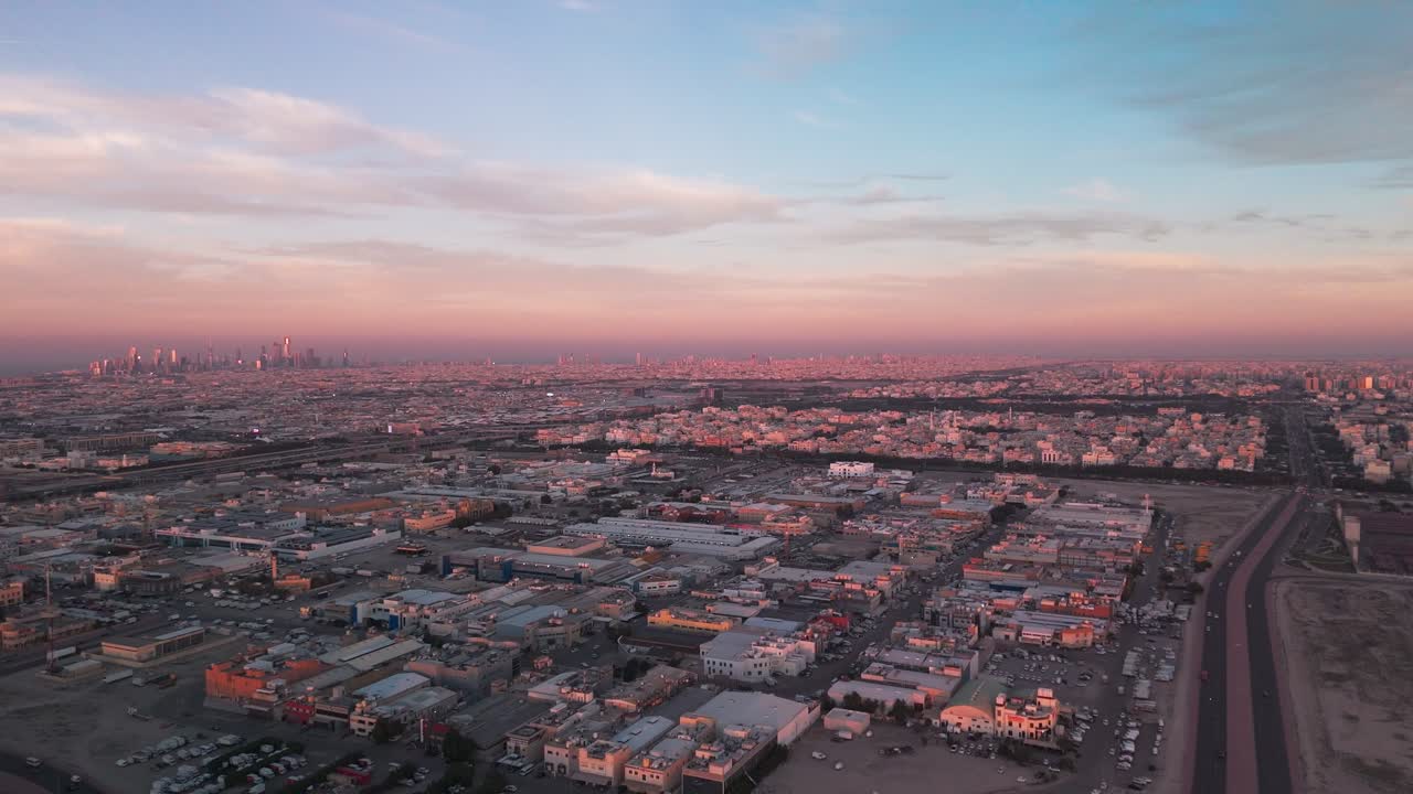 Ampia ripresa aerea con drone durante l'ora d'oro che mostra l'espansione urbana, i tetti industriali e il traffico autostradale che porta a un lontano skyline cittadino sotto nuvole pastello