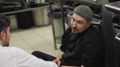 Communication-between-a-professional-cook-in-a-black-uniform-and-a-waiter-during-a-short-break-in-the-kitchen-while-sitting-on-the-floor-leaning-on-the-table-during-a-busy-working-day