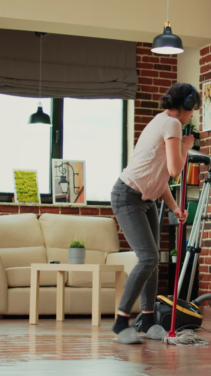 Vertical video: Happy housewife listening to music on headset and cleaning apartment