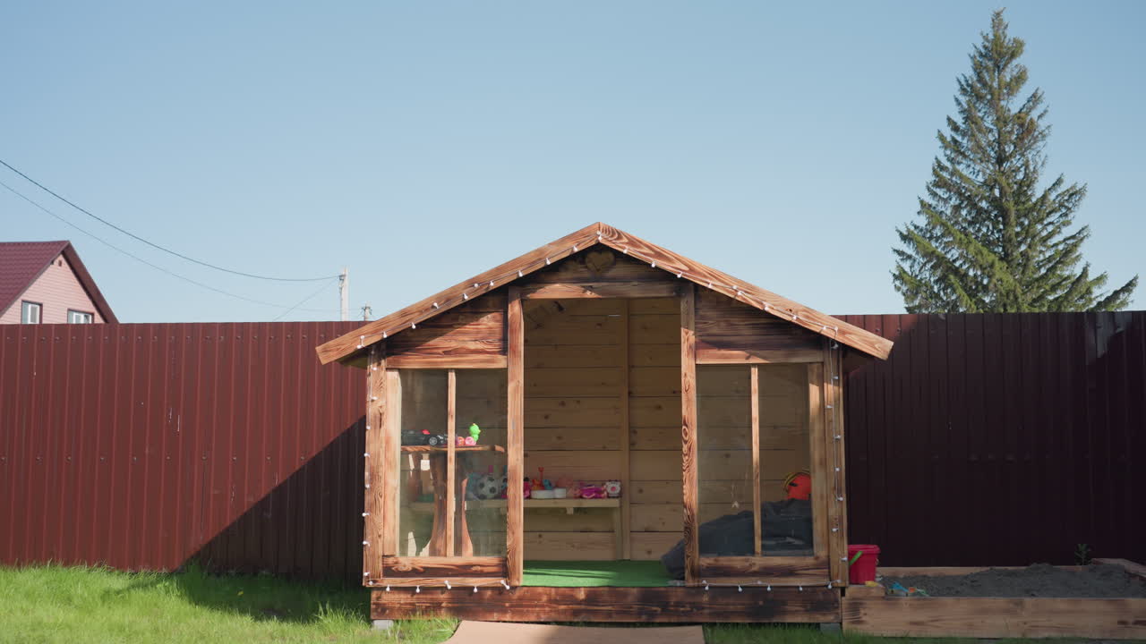 Wooden children playhouse with glass panels reflecting person filming, resting on green grass with small sandbox beside it, red fence and tall pine tree in background under clear blue sky